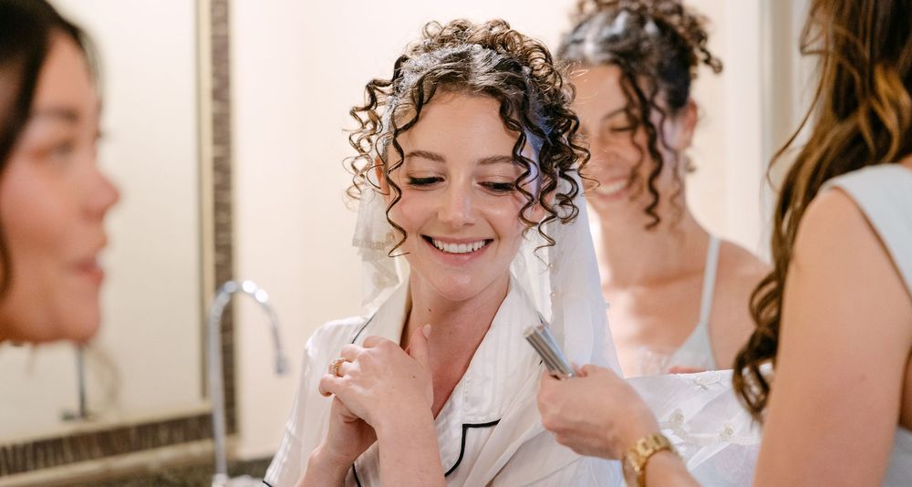 A person with curly hair smiles joyfully while getting ready in a white button-up shirt.