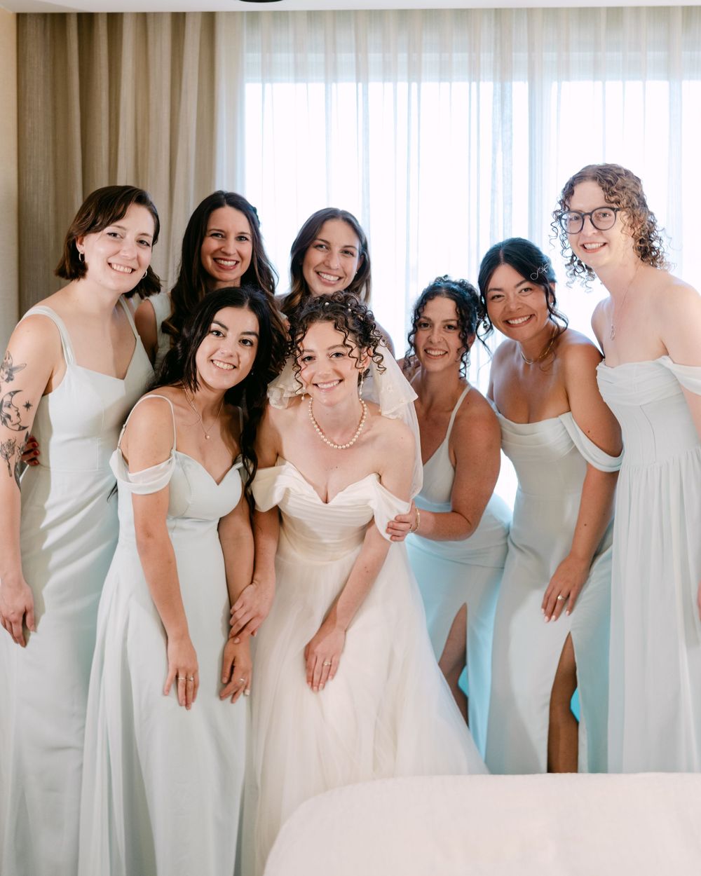 A bridal party poses together wearing pastel blue bridesmaid dresses and a white wedding gown in a sunlit room.