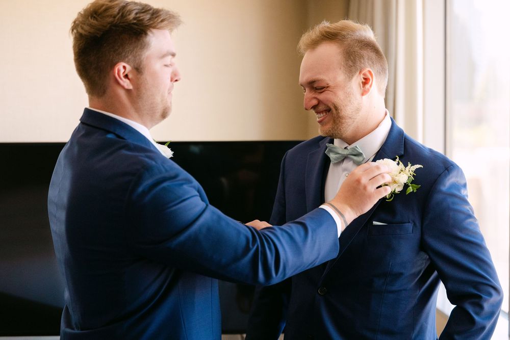 Two people in navy blue suits adjust boutonnieres and share a joyful moment before a wedding ceremony.