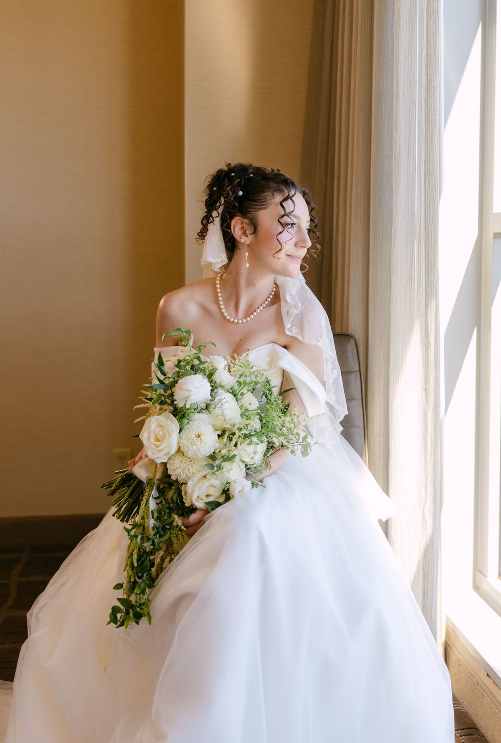 A bride in a white ballgown wedding dress stands by a window holding a cascading bouquet of white roses and greenery.