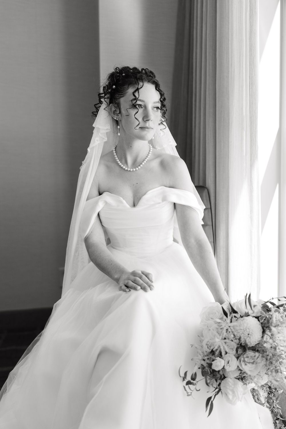 A bride in an elegant off-shoulder white gown poses by a window with her bouquet in black and white photography.