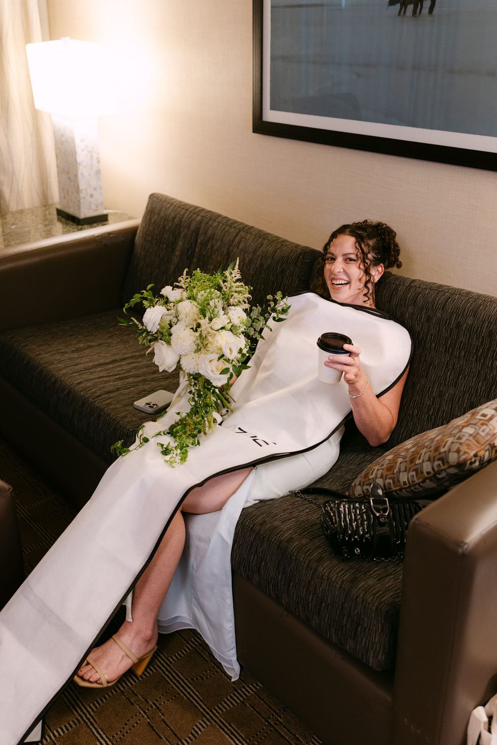 Someone in an elegant white dress relaxes on a dark couch with a bouquet of white flowers and greenery nearby.