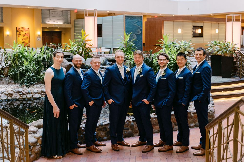 Wedding party in navy suits and formal attire pose together in an elegant indoor venue with wooden railings.