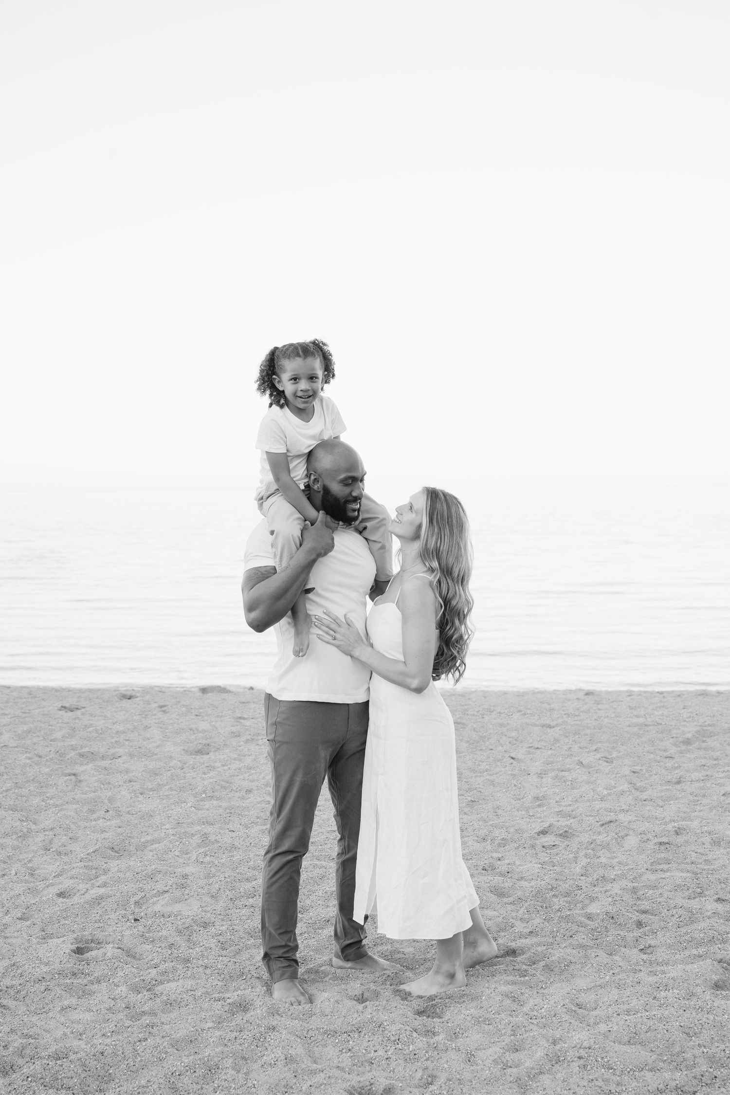 A couple in white outfits embracing and dancing together on a sandy beach in black and white. - Lake Forest