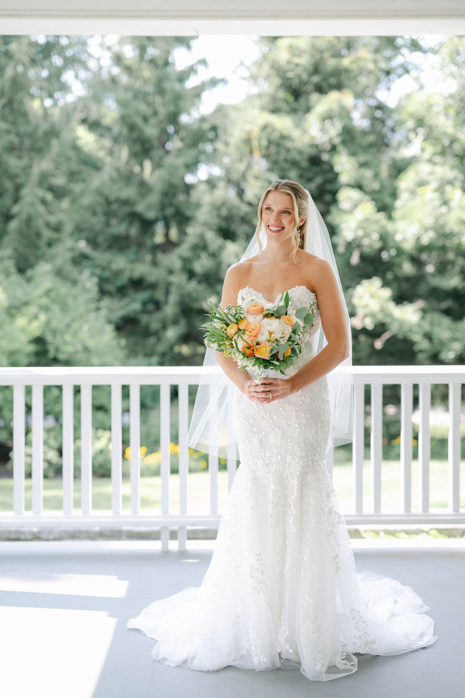 A bride in a white lace wedding dress poses elegantly on a classic white columned porch with a colorful floral bouquet. - Lake Forest