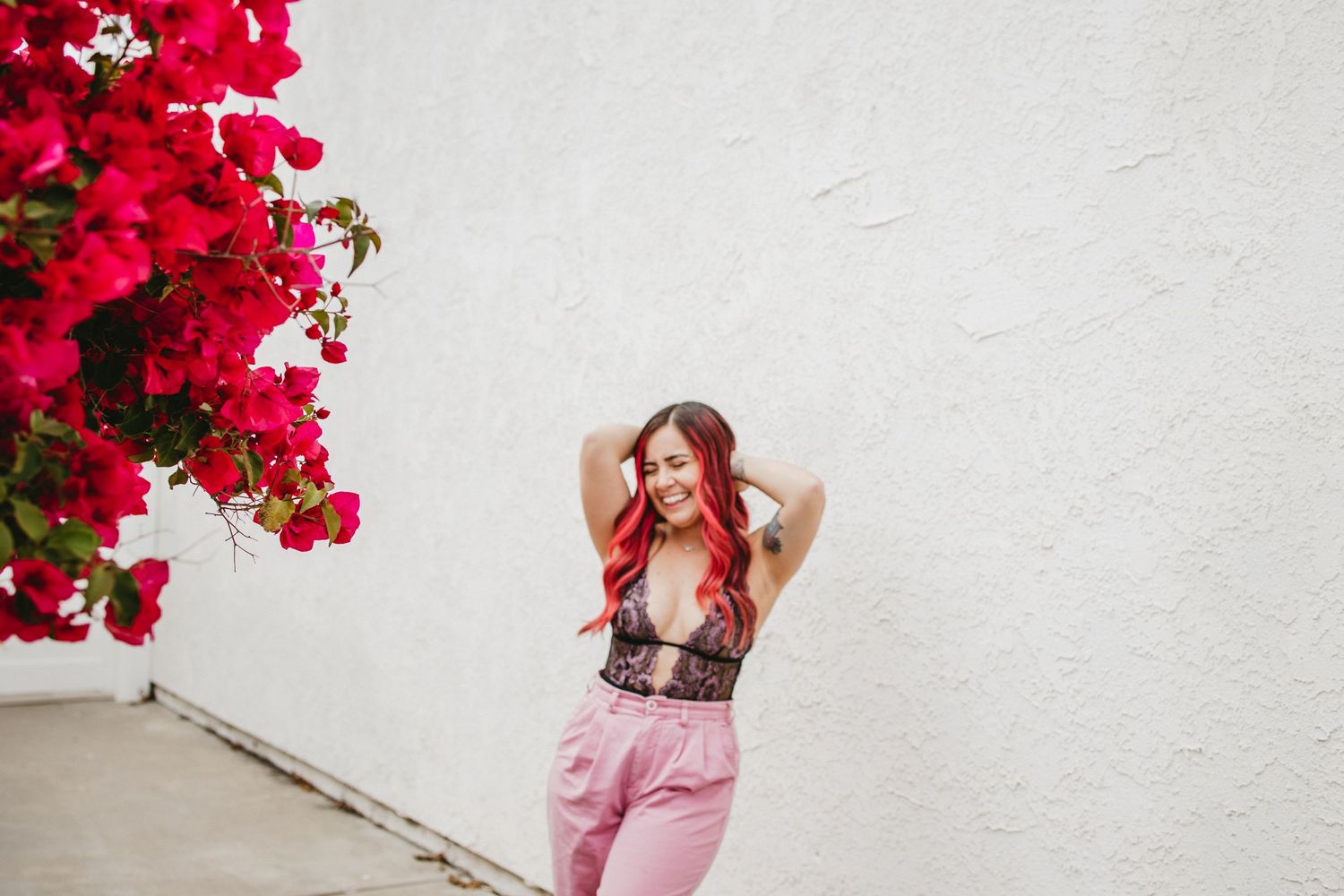 Woman with pink hair, wearing a black and pink lingerie body suit, with pink pants, next to vibrant bougainvillea plant
