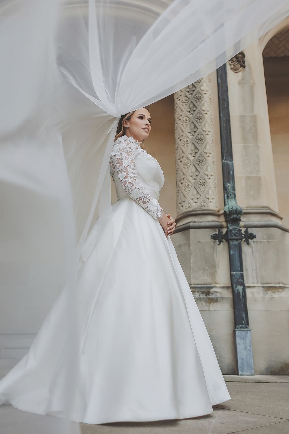 Elegant white wedding gown with lace sleeves and flowing veil captured in a dramatic architectural setting.