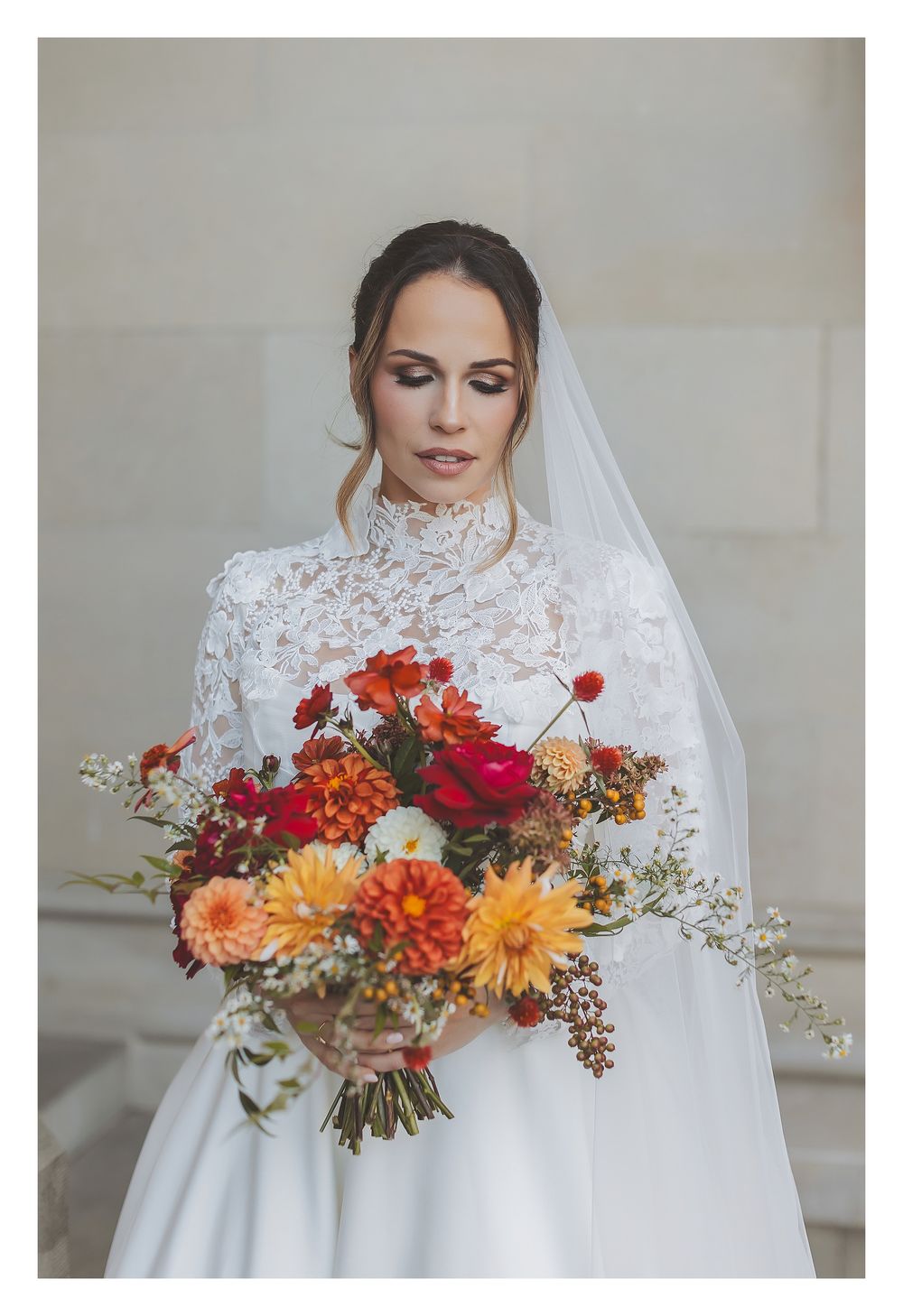Bride in white lace wedding dress holding vibrant autumn bouquet with orange dahlias, red roses and delicate foliage.