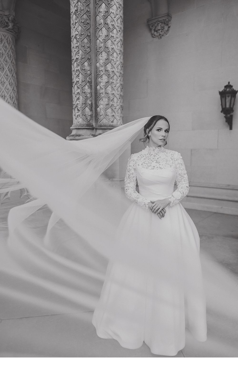 A bride in a classic long-sleeved wedding gown poses elegantly on marble steps in black and white photography.