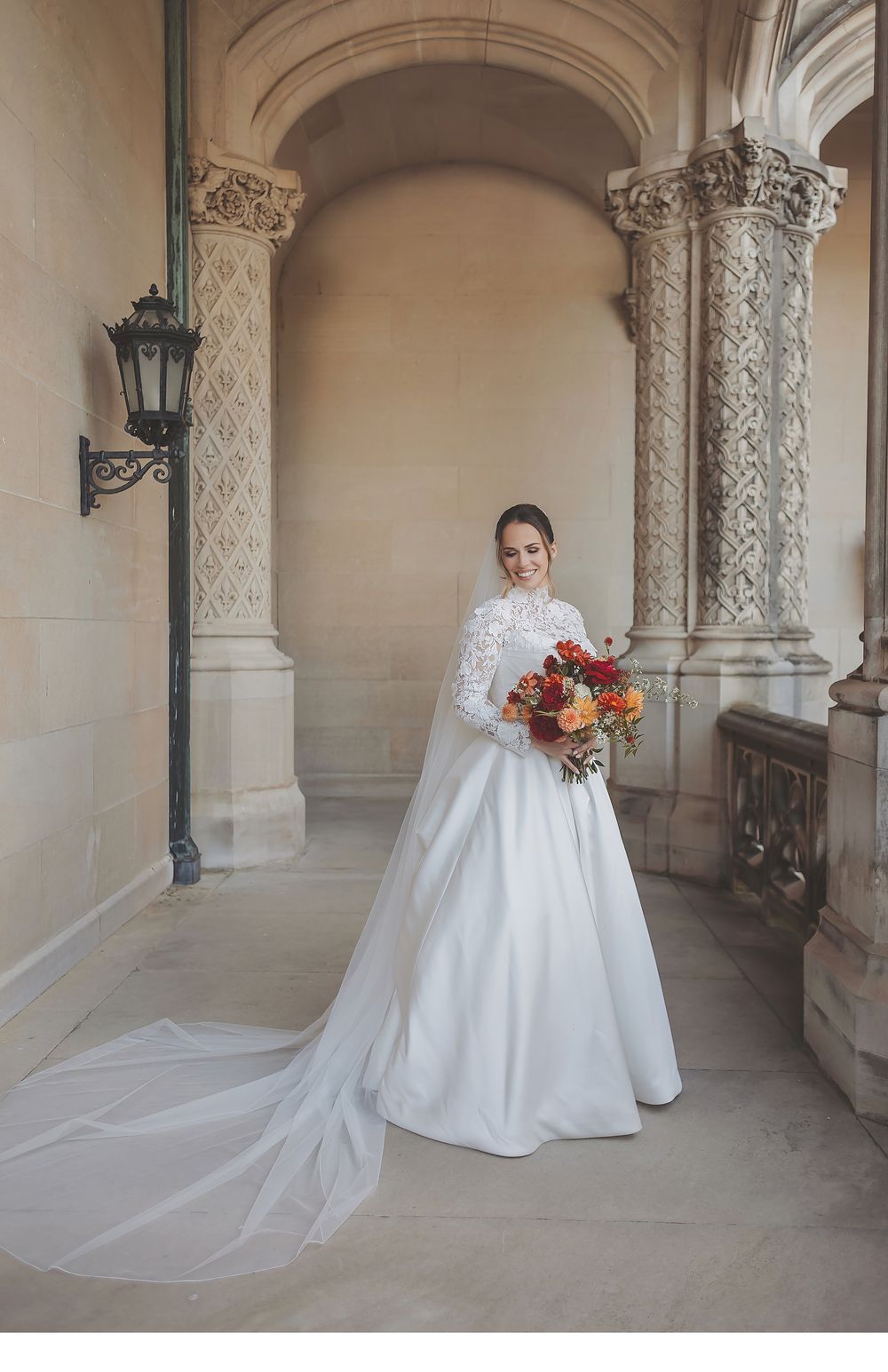 A bride in a classic white wedding gown with a flowing train poses in an elegant stone archway corridor.