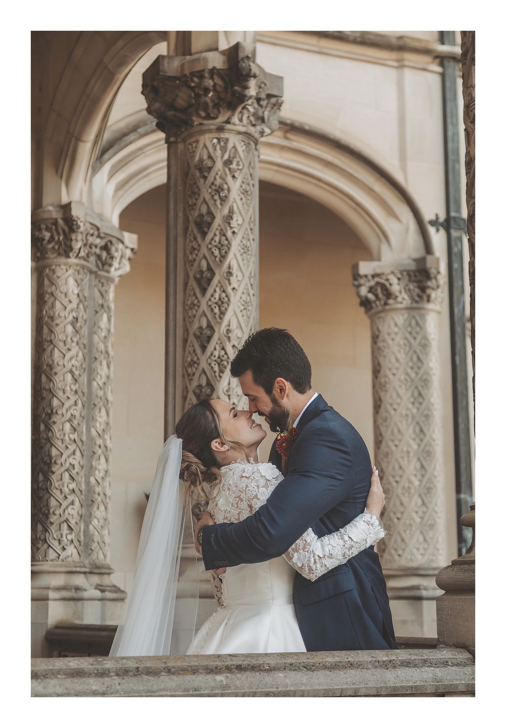 A romantic wedding couple shares a tender kiss under ornate stone archways with decorative columns at a historic venue.
