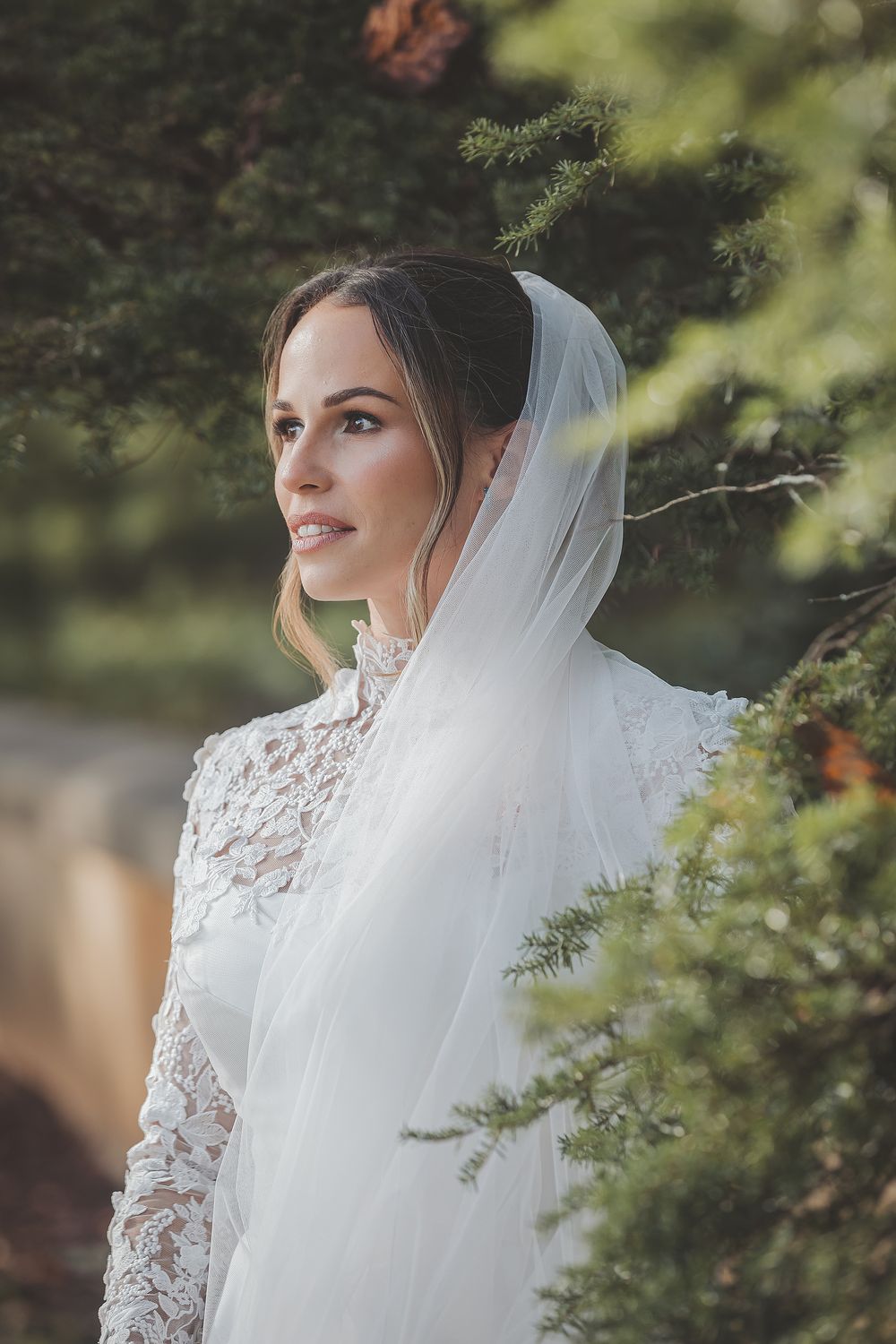 A bride in a long-sleeved lace wedding gown and veil poses elegantly against a natural green outdoor background.
