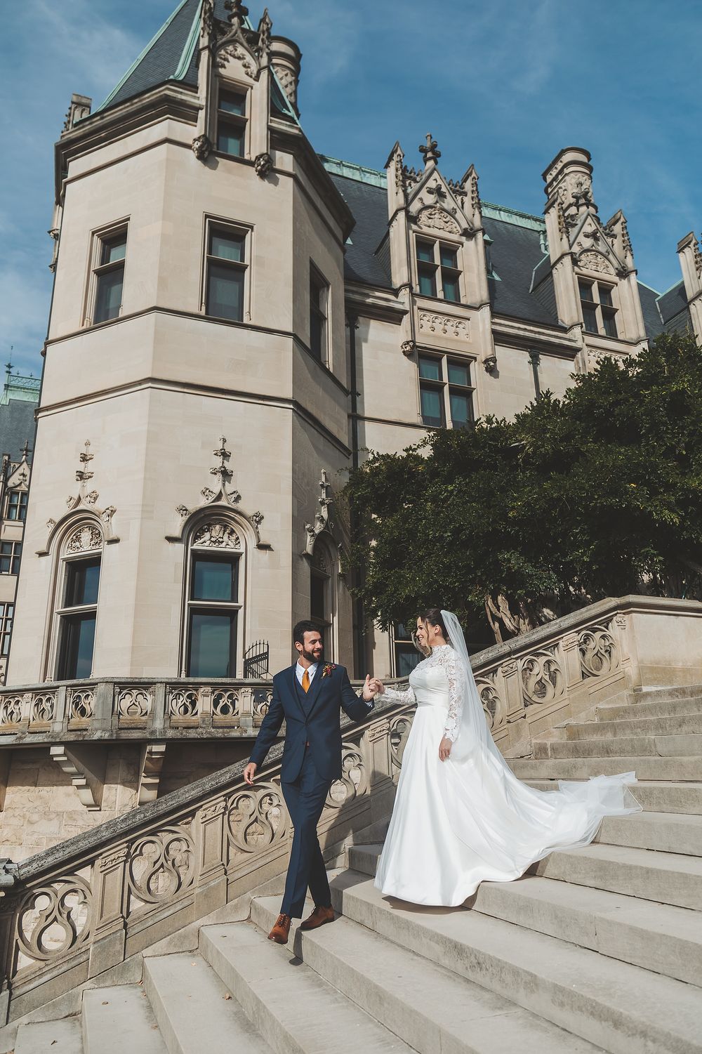 A couple in wedding attire poses on grand stone steps of an elegant historic building with ornate architecture.