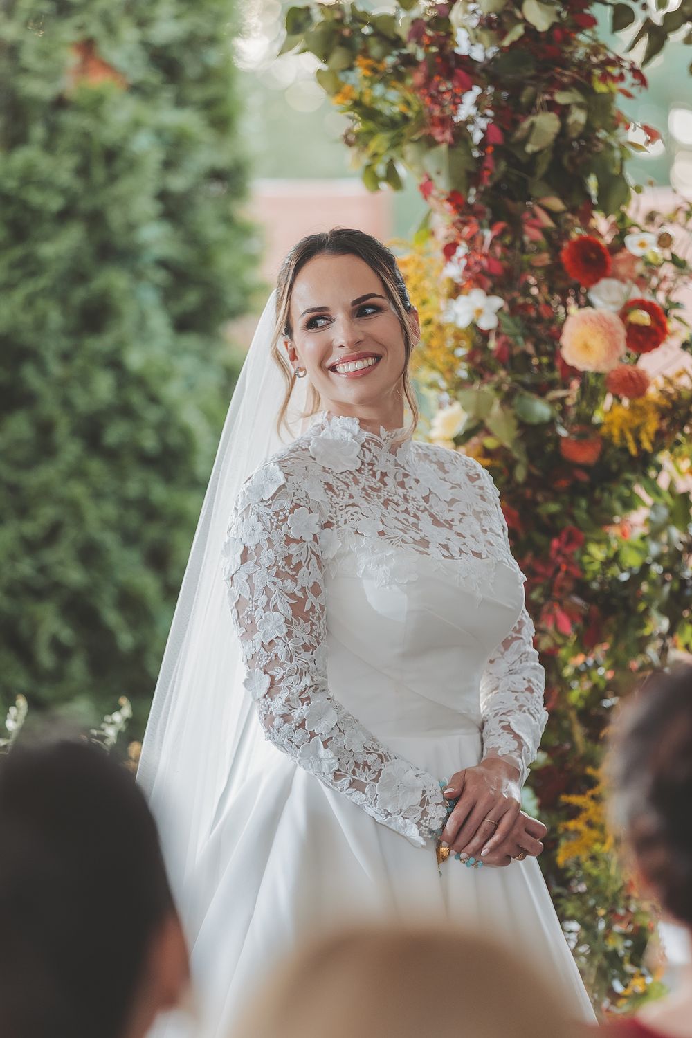A bride in a long-sleeved lace wedding dress stands under a floral arch during an outdoor ceremony.
