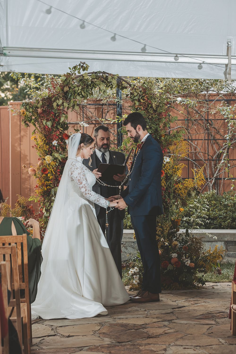 A wedding ceremony takes place in a garden courtyard with string lights and autumn foliage decorations.