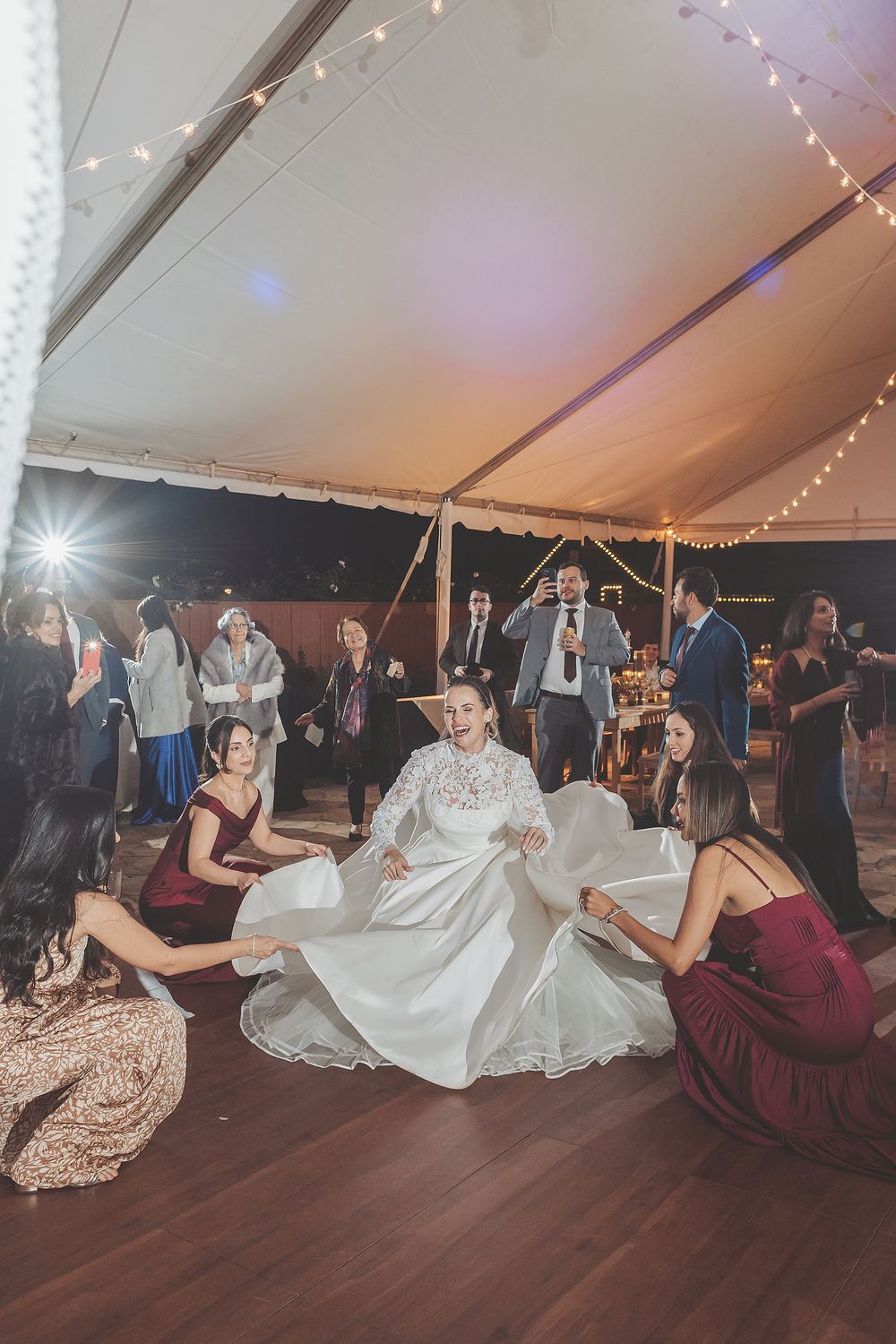 People dancing in a line formation at a wedding reception under a tent with twinkling lights overhead.