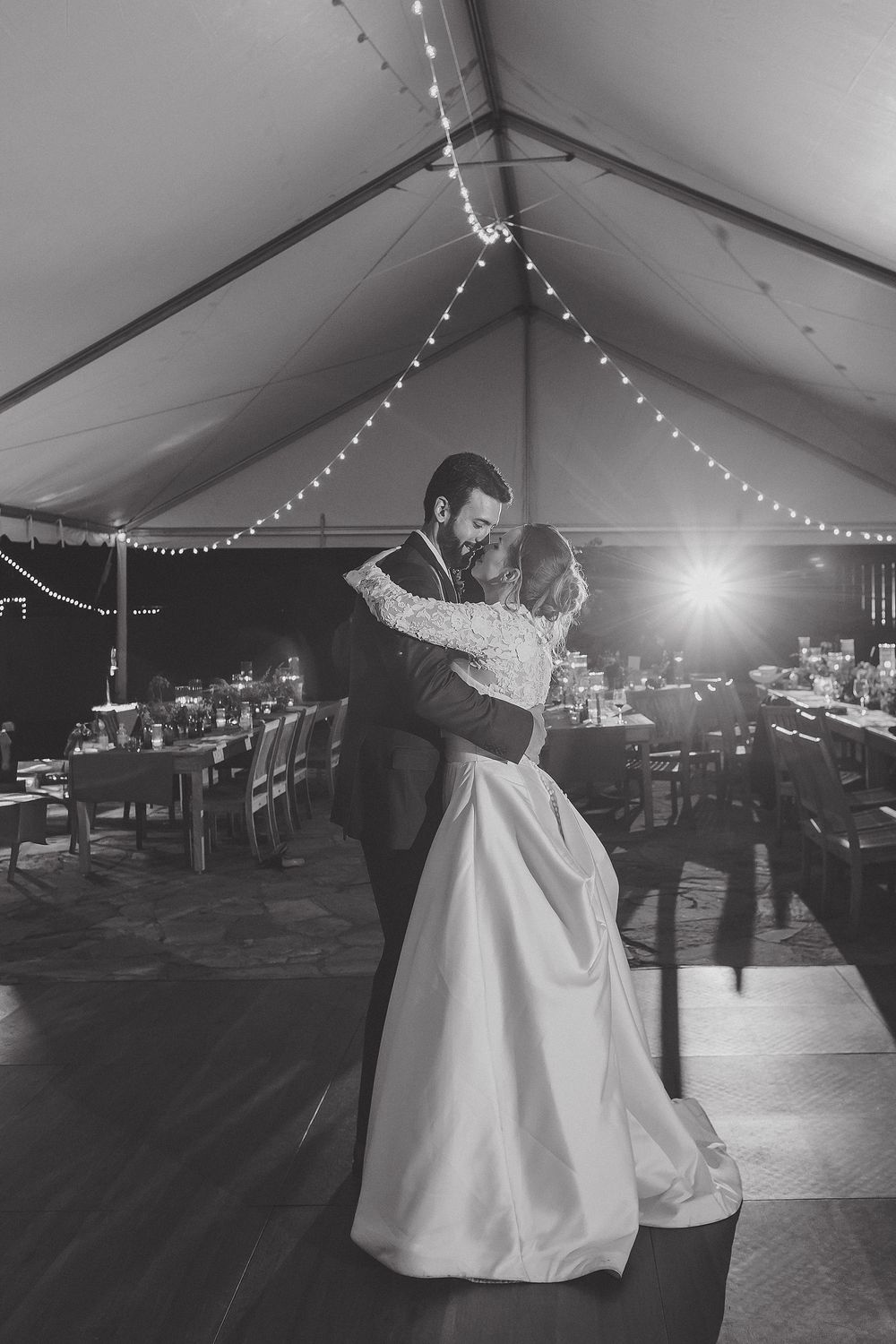 A couple shares their first dance under string lights in a wedding reception tent with tables in the background.