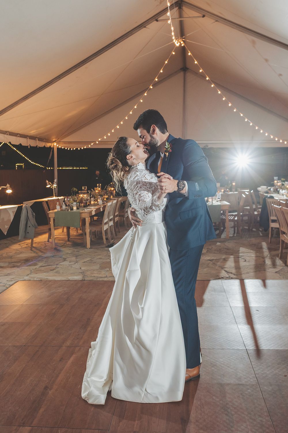 A couple shares their first dance under twinkling lights in a rustic tent reception venue with wooden floors.