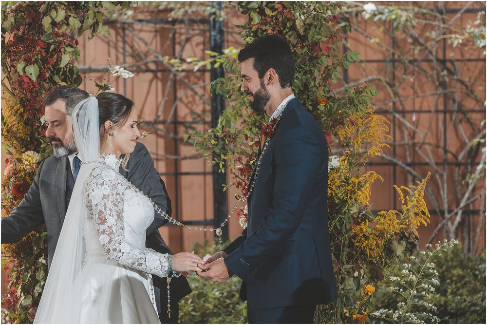 A wedding ceremony takes place in a garden setting with brick walls and autumn foliage in the background.