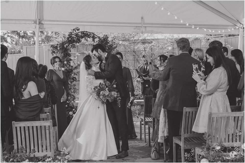 A black and white wedding ceremony photo showing a couple at the altar under a tent with guests looking on from their seats.