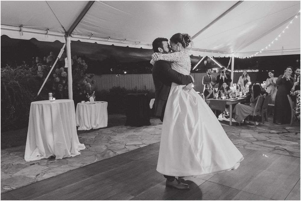 A couple shares a romantic dance under a white tent at an outdoor wedding reception during sunset.