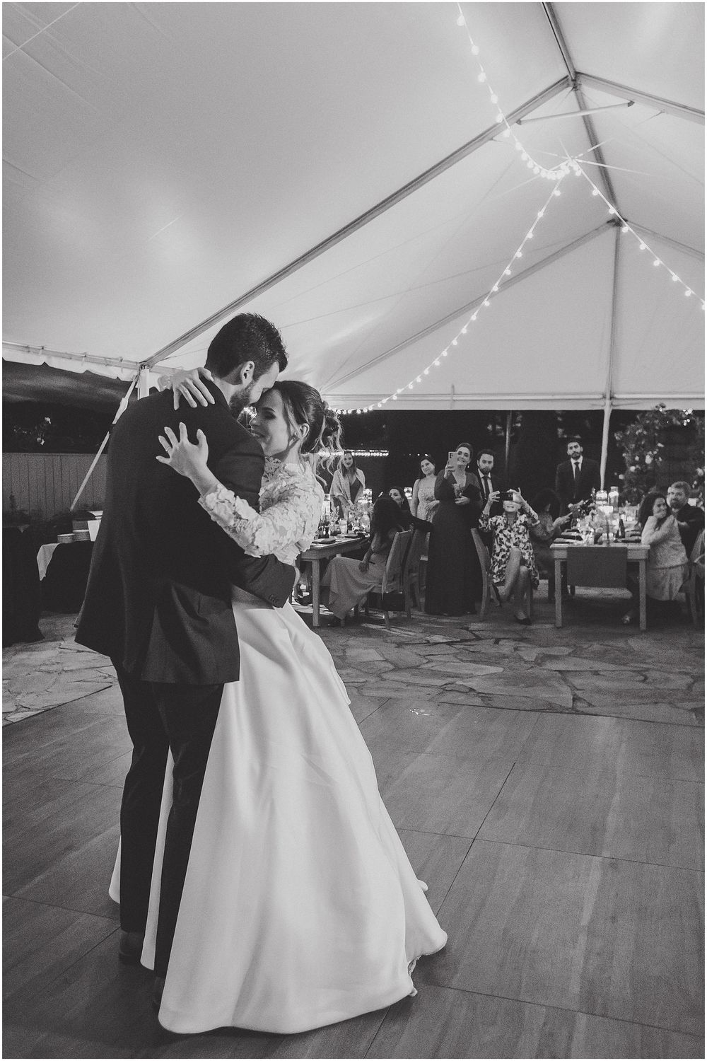 A couple shares their first dance under a white tent at an elegant outdoor wedding reception with string lights overhead.