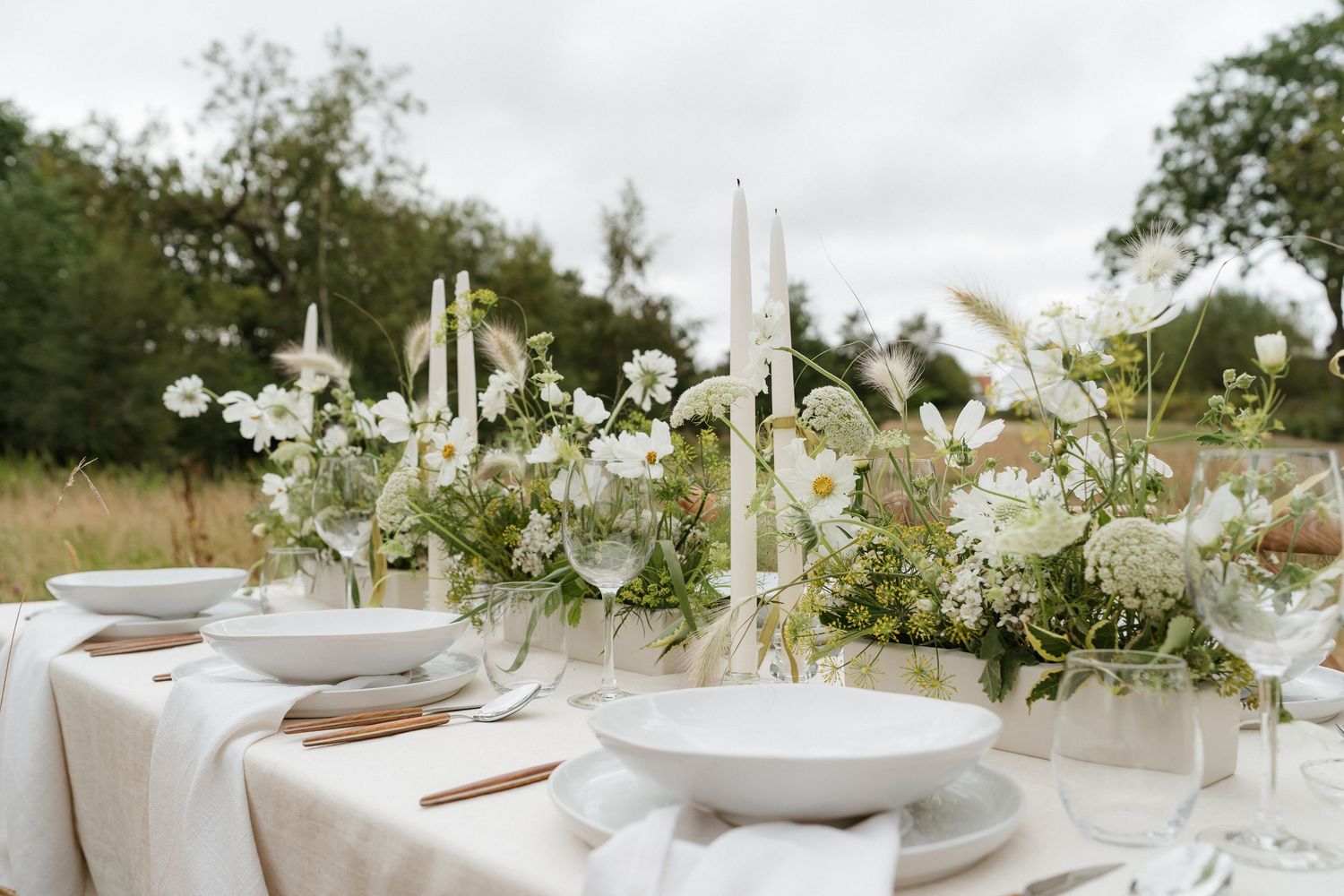 Elegant outdoor table setting with white linens, candles, and floral arrangements in a rustic garden setting.
