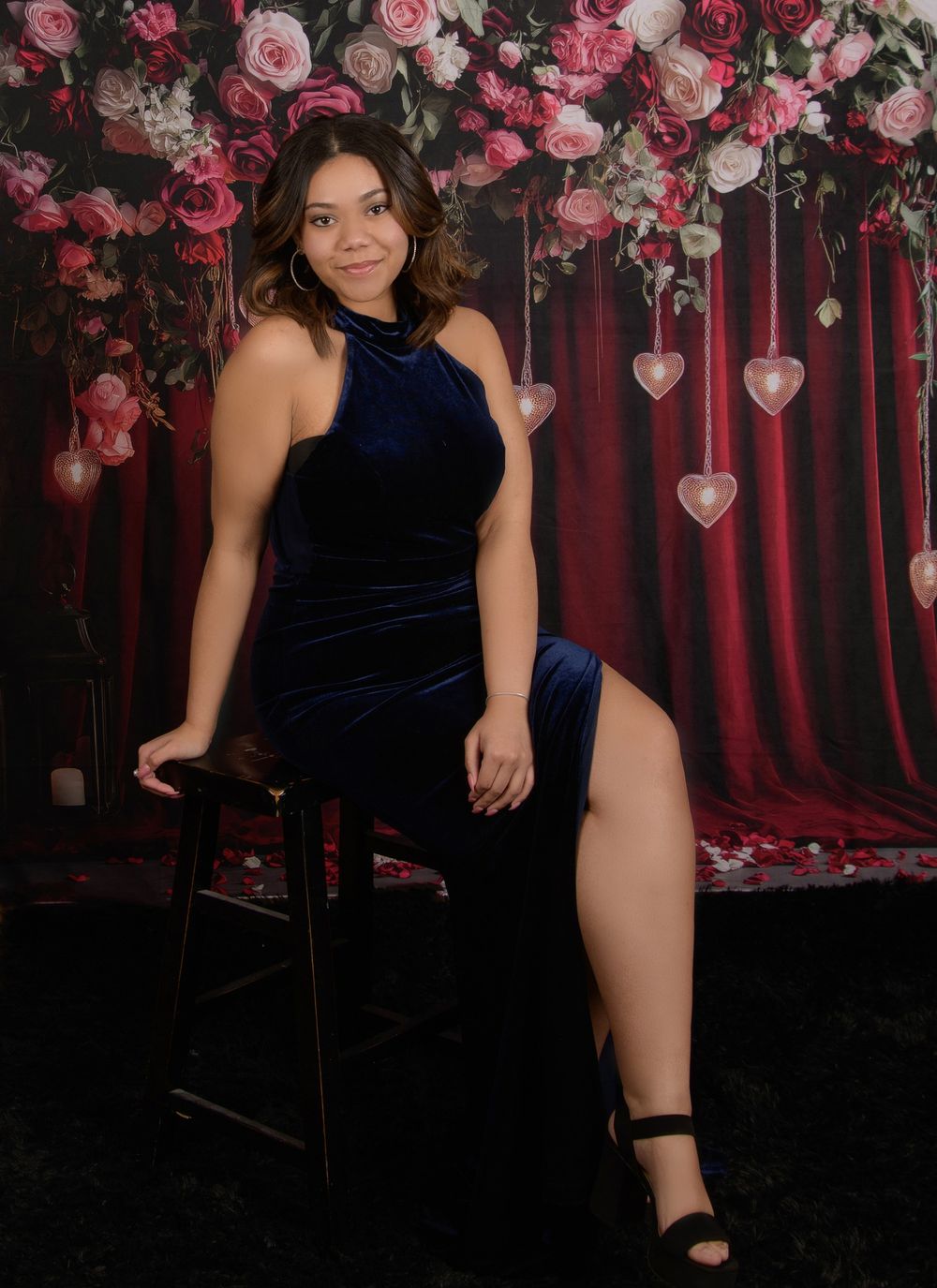 Romantic photo shoot setup with pink roses and hanging heart decorations against a dark red curtain backdrop.