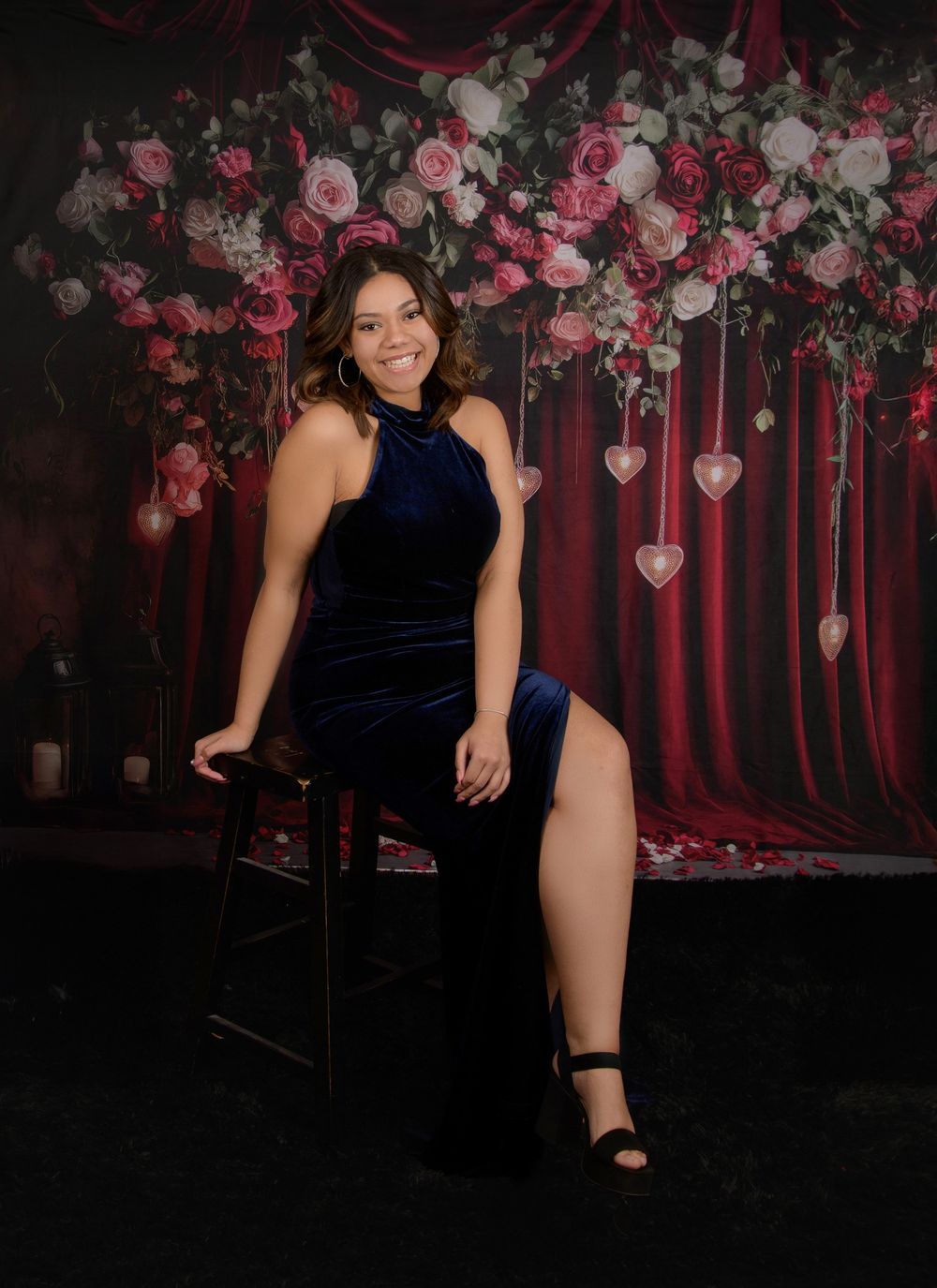 A model in a navy velvet bodysuit poses against a romantic backdrop of pink roses and red drapery.