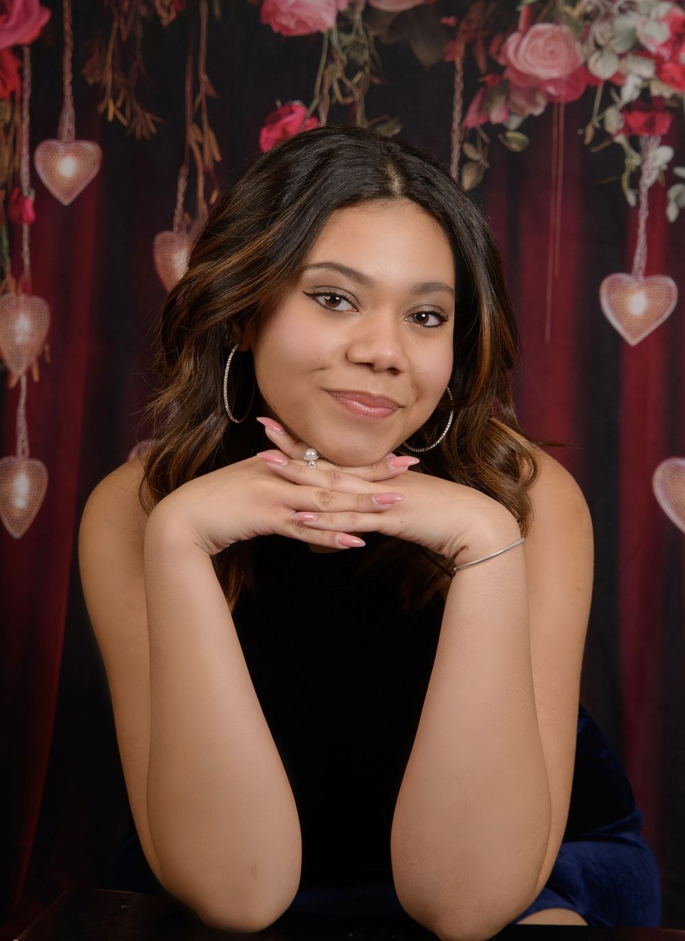 A person in a dark blue top poses with hands under chin against a romantic backdrop with pink heart decorations.