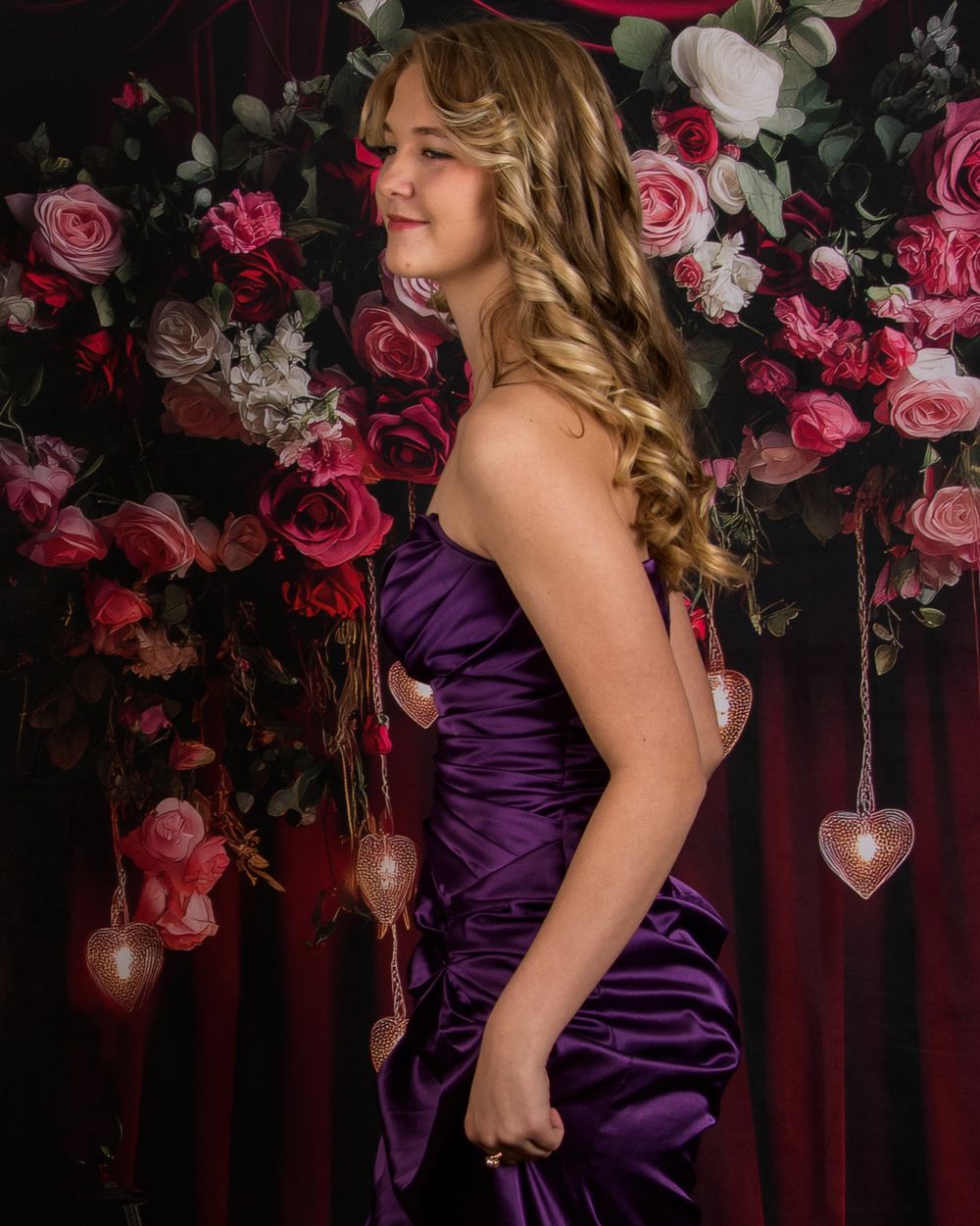 Woman in elegant purple satin gown poses against dark backdrop with pink and white floral arrangements and hanging lights.