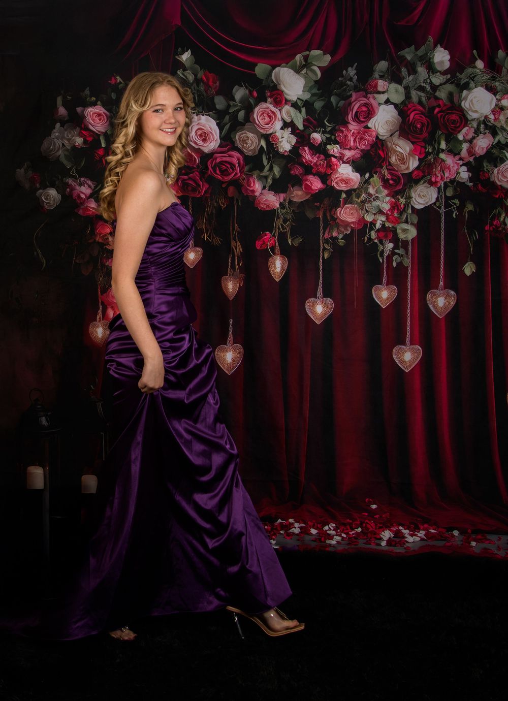 A person in a purple satin gown poses against a dark backdrop with pink and white roses and hanging heart decorations.