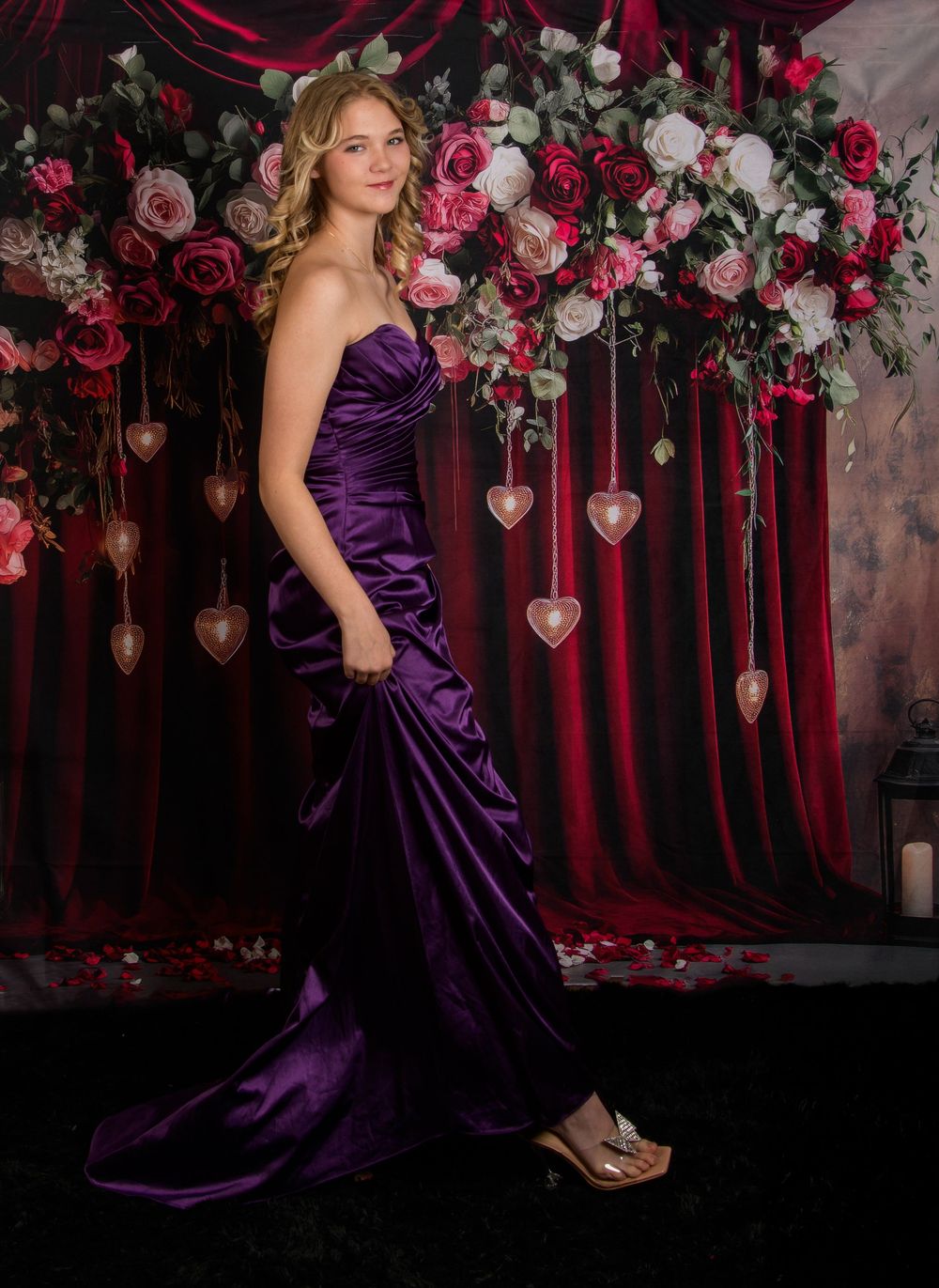 Elegant model in deep purple satin gown poses against romantic backdrop of roses and red velvet curtains.