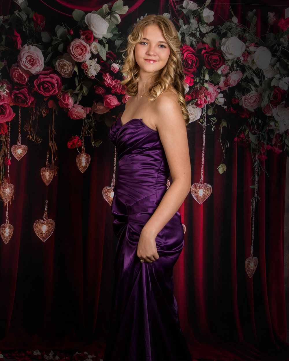 A figure in a purple satin gown poses elegantly against a romantic backdrop of pink roses and hanging heart decorations.