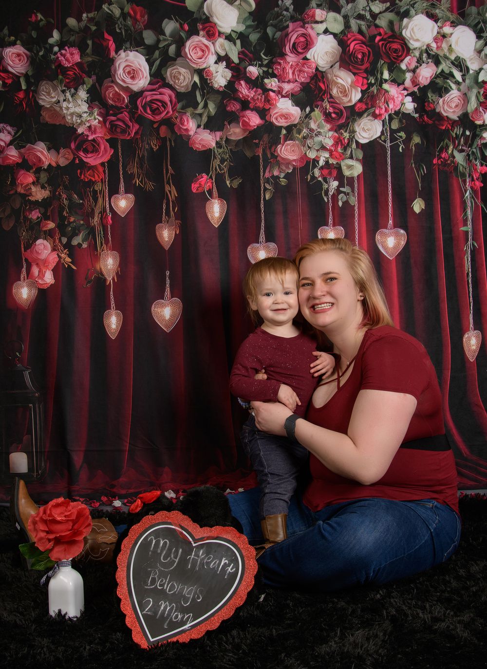 Valentine's day photo setup with hanging pink roses, white flowers, and heart decorations against a dark red backdrop.