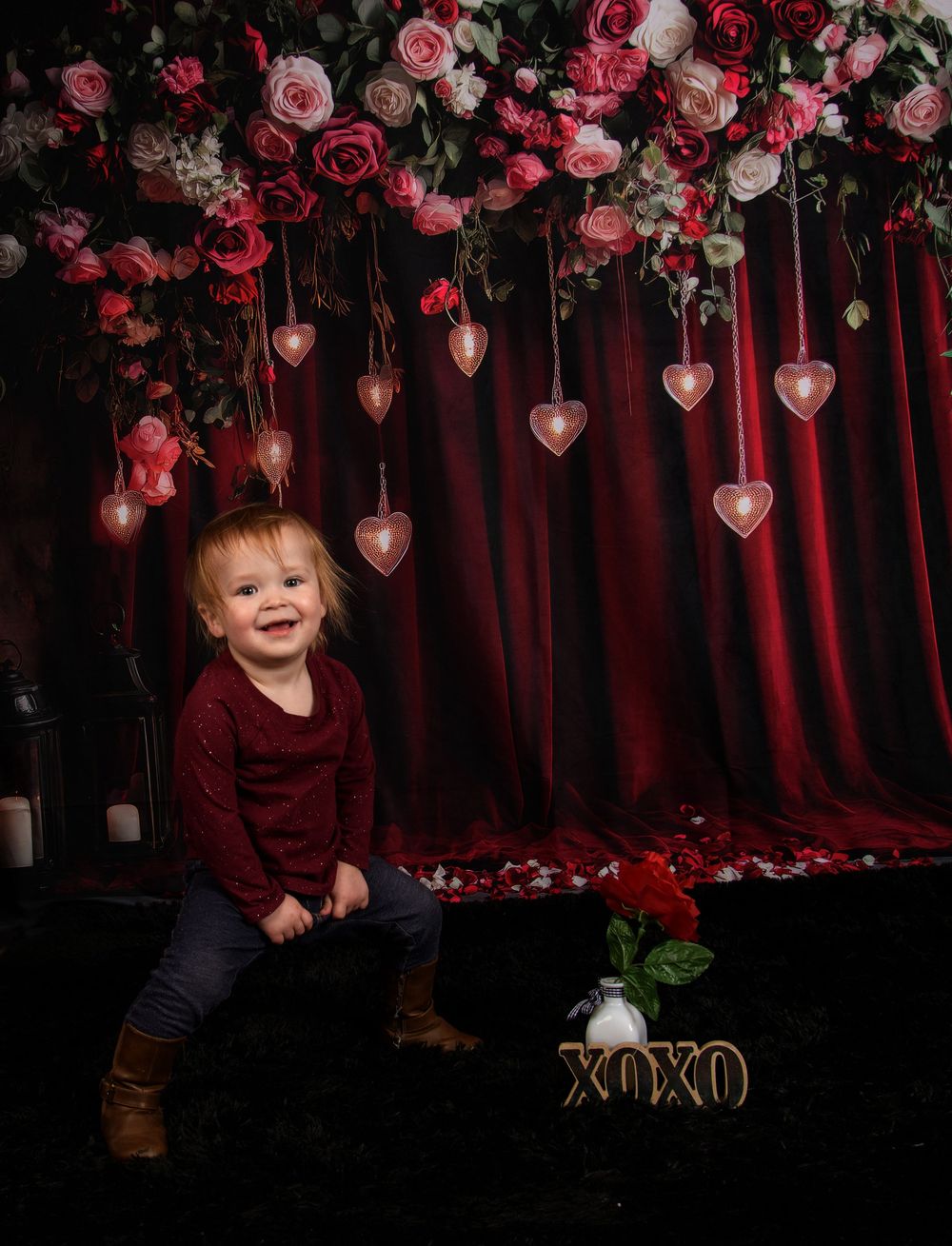A romantic Valentine's Day photo setup with hanging heart decorations, pink roses, and burgundy curtain backdrop.