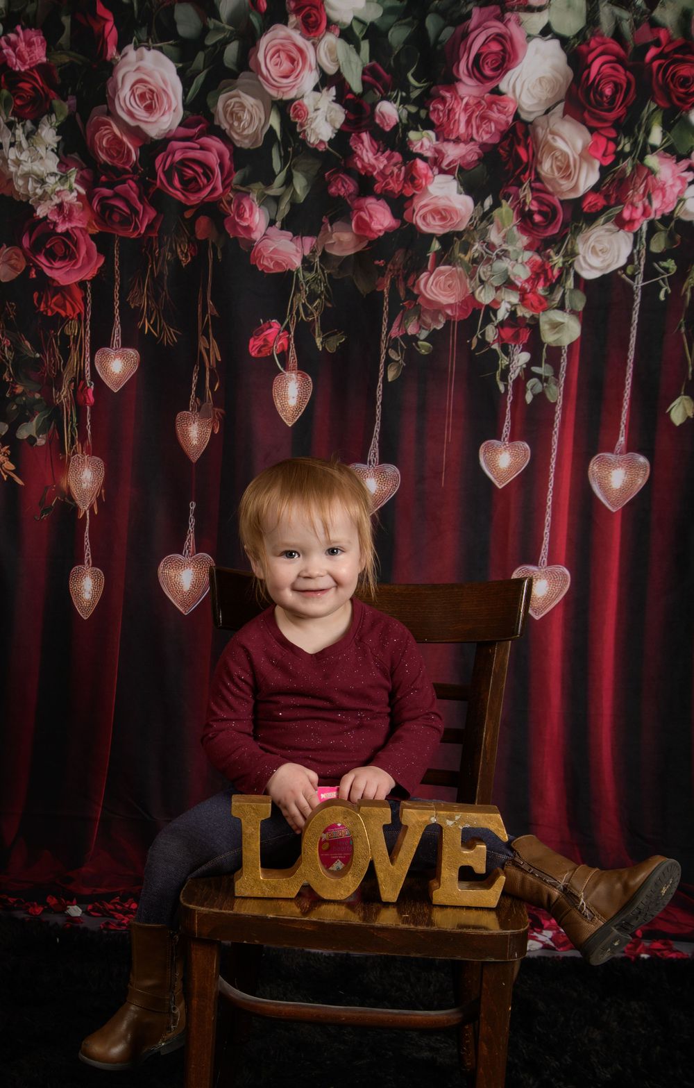 Romantic Valentine's photo setup with pink roses, hanging heart decorations, and a wooden bench against burgundy curtains.