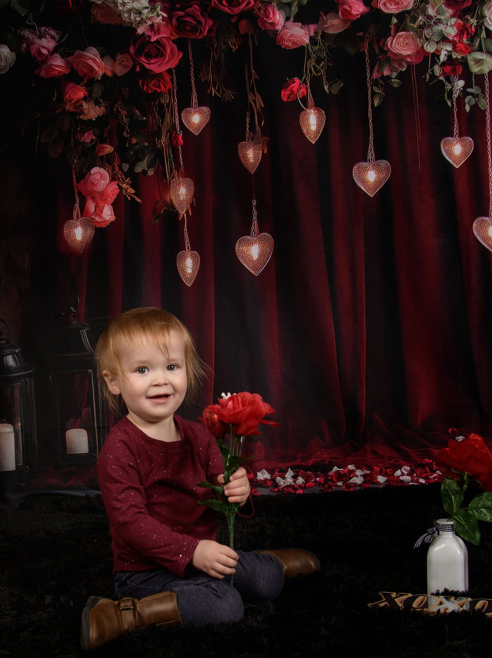 A romantic Valentine's Day photo setup with hanging heart decorations, red roses, and burgundy backdrop.