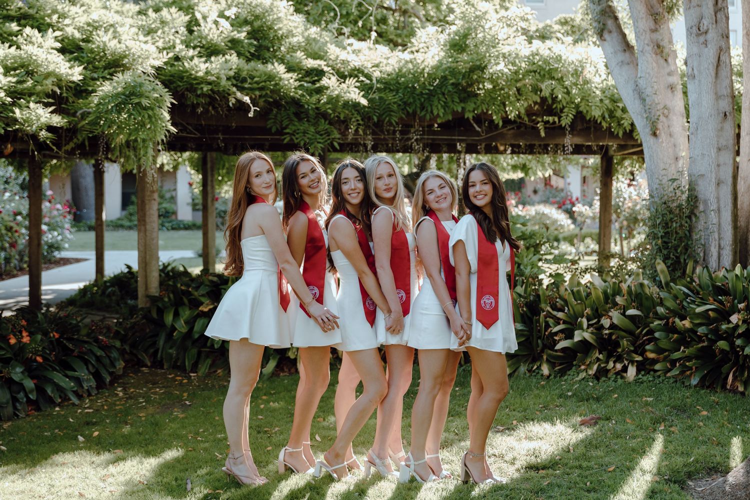 Group of girls wearing red sashes and white dresses poses together at Santa Clara University for their group graduation photoshoot.