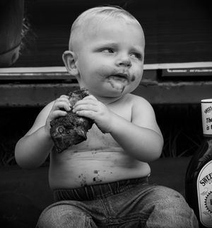 A young child in overalls sits and eats while looking off into the distance in black and white.