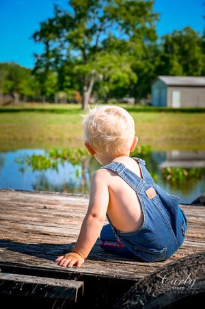 Child in denim overalls sits peacefully on wooden dock overlooking a serene pond.