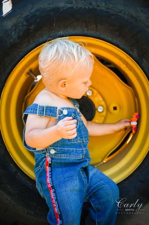 Toddler in overalls examines large yellow tractor wheel during country photoshoot.