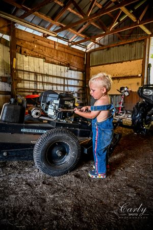Small child in overalls explores vintage farm equipment inside a rustic wooden barn.
