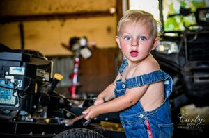 A young child in denim overalls stands at a workbench in a garage workshop setting.