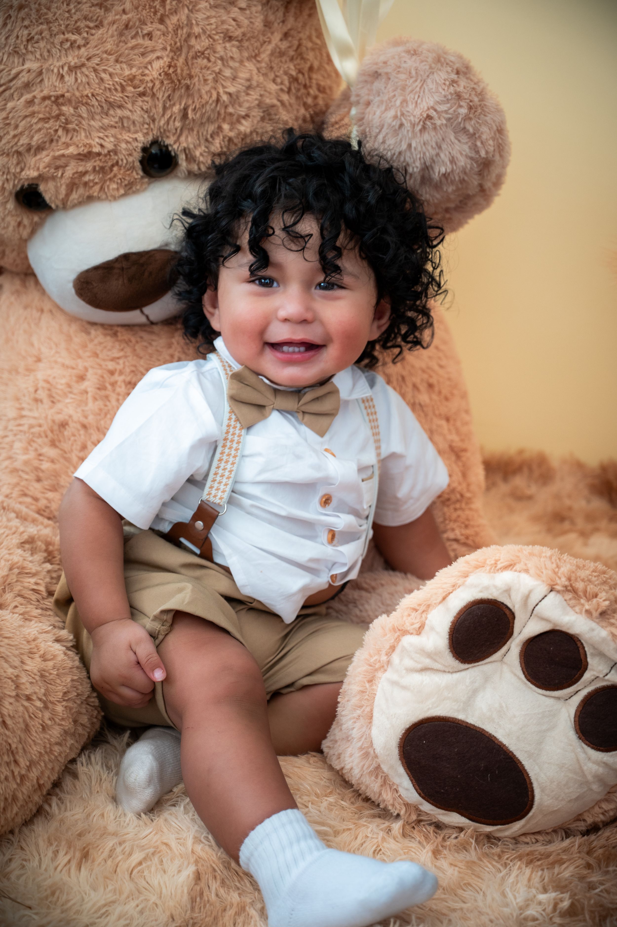 Toddler in white shirt and khaki shorts sitting on large plush teddy bear with paw visible in corner.