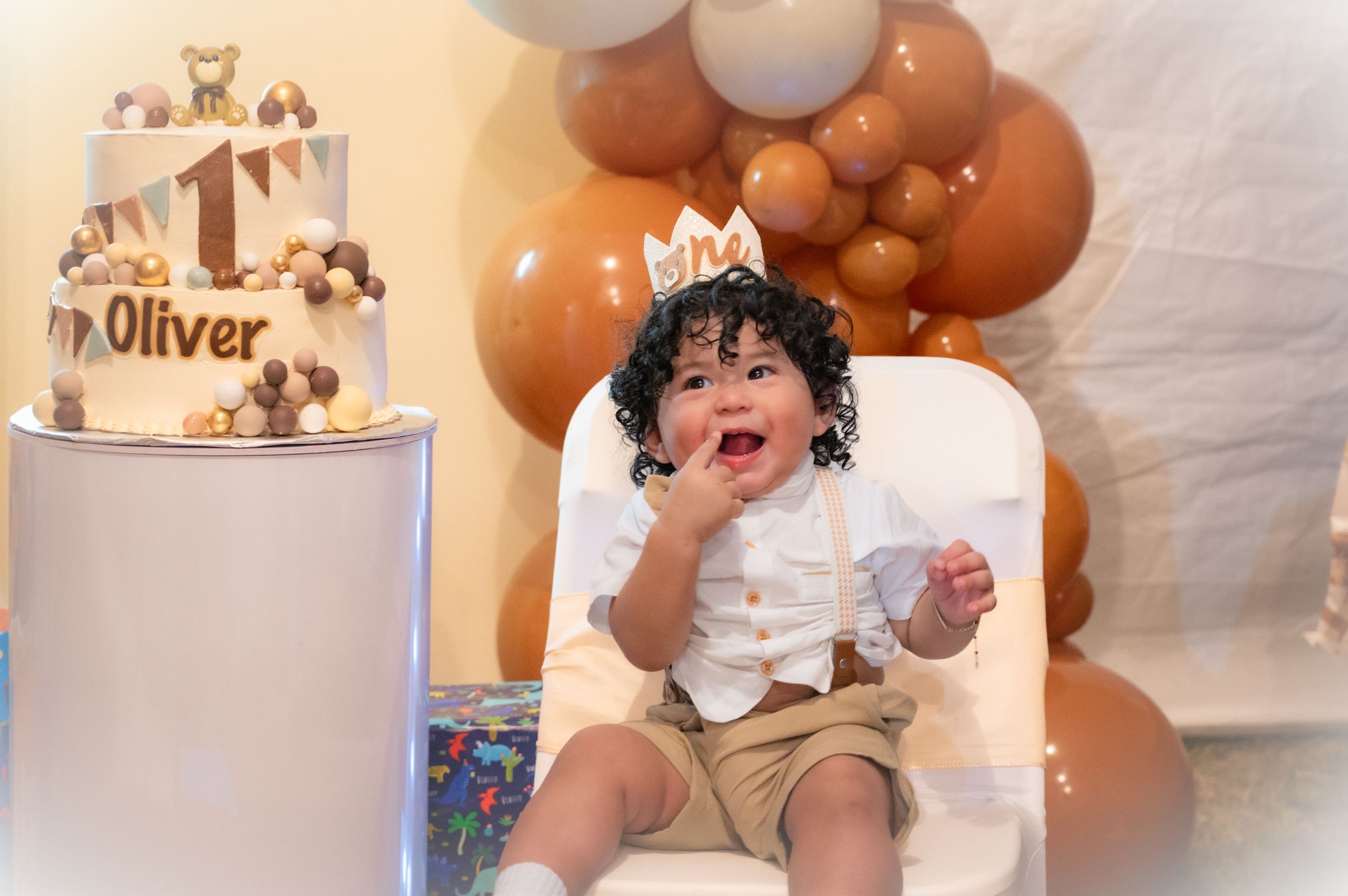 A decorated birthday party setup with a white cake, brown balloons, and a small child sitting in a chair.