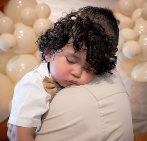 A child with curly dark hair rests peacefully against a shoulder in a white shirt with beige balloons in the background.