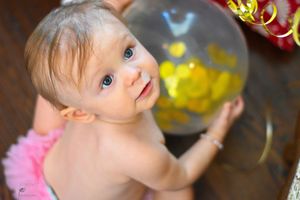 Baby in pink tutu reaching up to touch a large balloon with yellow decorations inside.