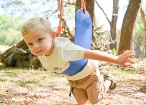 A toddler wearing a blue baseball cap plays outdoors in a wooded area on a sunny day.