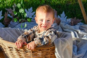 A child sits nestled in a wicker basket outdoors with greenery and trees visible in the background.
